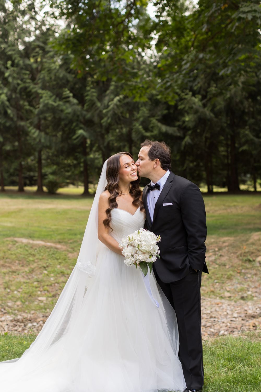 Groom in black tuxedo and bow tie kisses the cheek of bride in strapless gown with a long veil, holding a white bouquet outdoors.