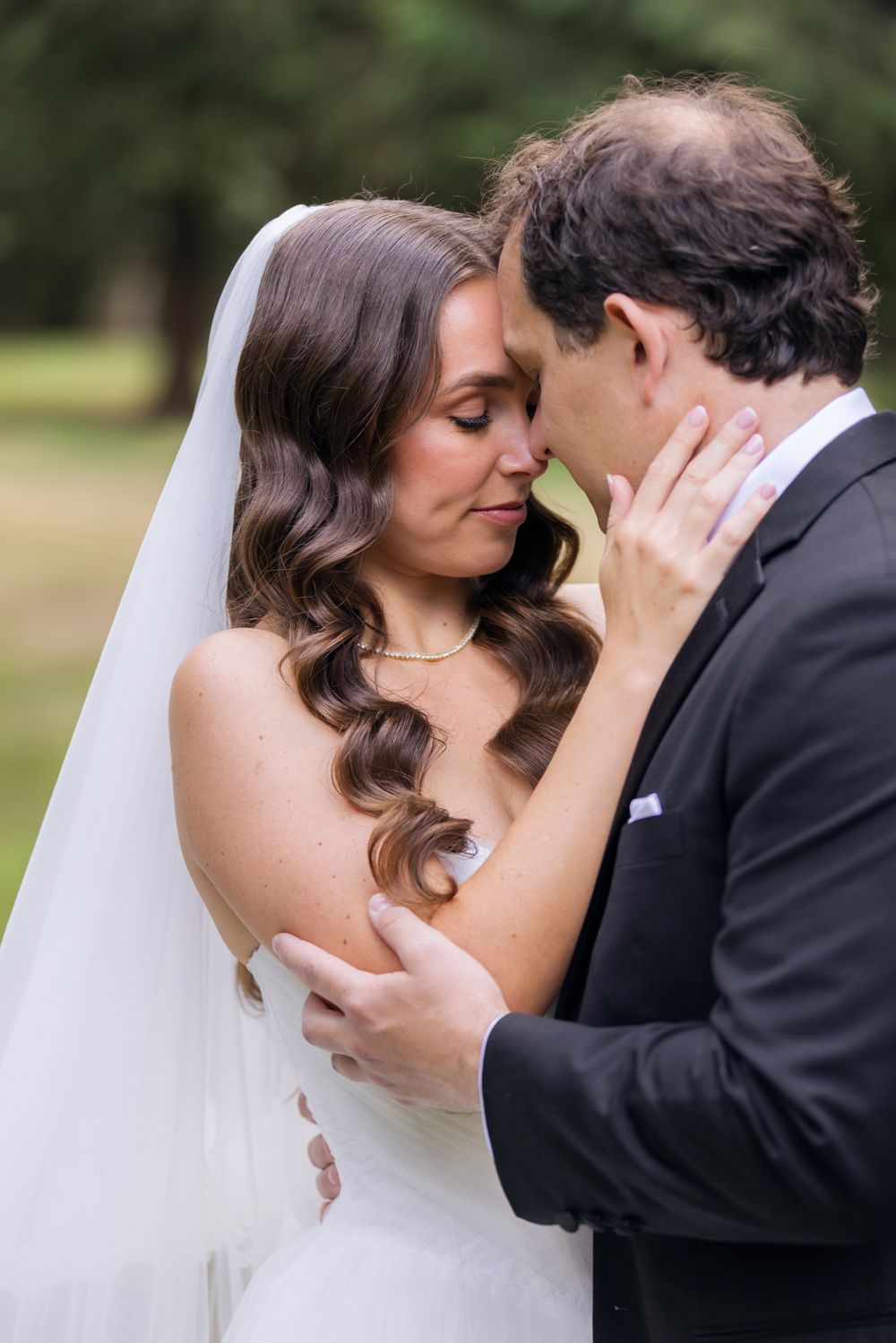 Bride in white gown and long veil and groom in black touch foreheads during tender outdoor wedding portrait. Groton wedding photographer