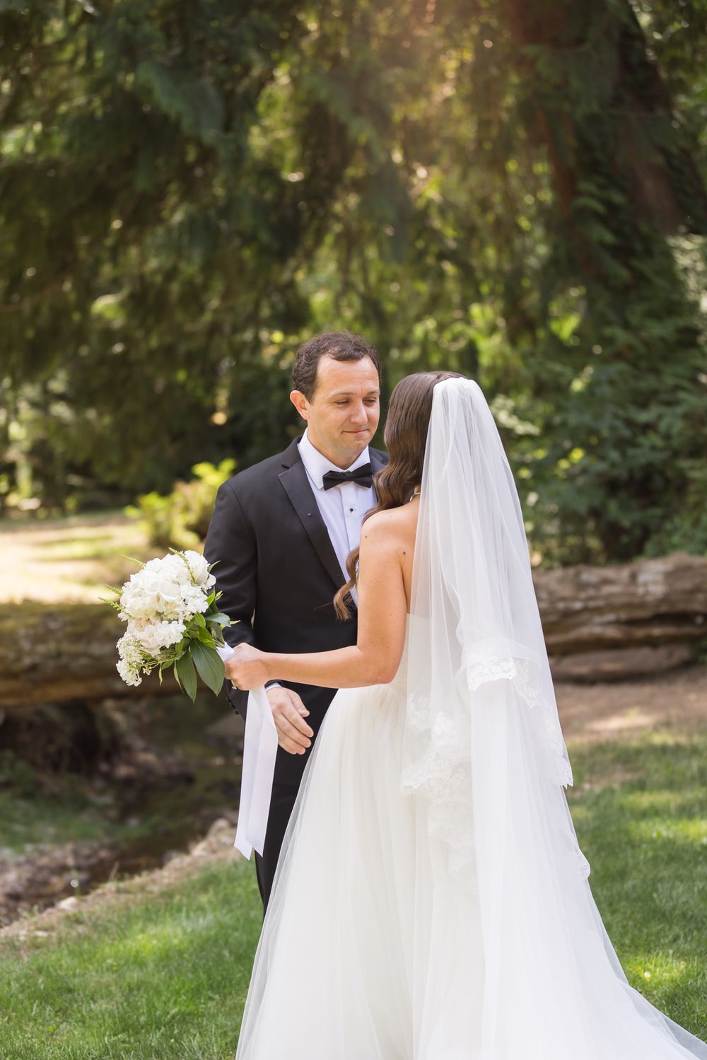Smiling groom in a black tuxedo and bow tie holds a white bouquet while facing a bride in a flowing gown and long veil for first look photos