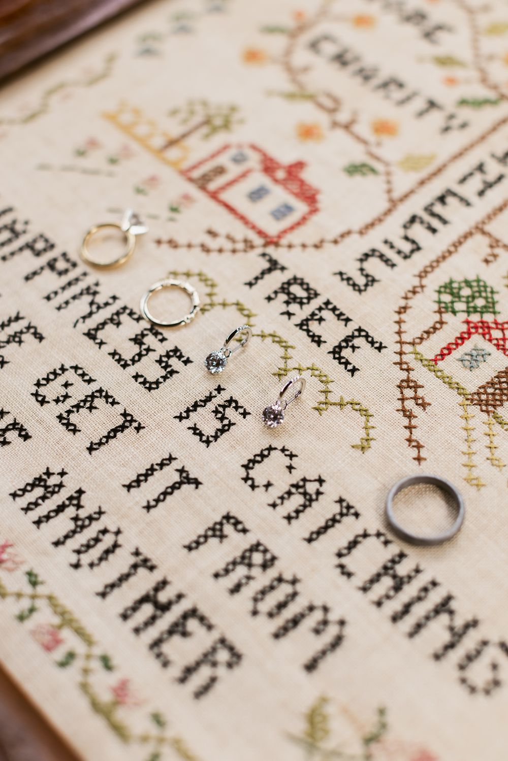 Wedding rings and diamond earrings rest on vintage sewing sampler stitched with a quote about happiness. North Shore wedding photographer