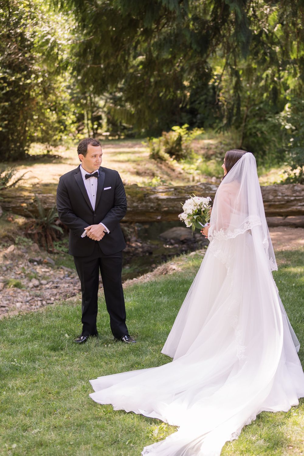 Groom gazes at bride with long veil as she approaches n forested setting with brook for first look photos. Concord wedding photographer