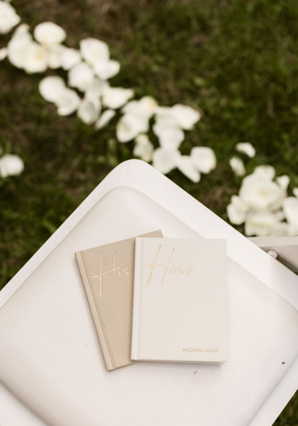 His and Hers wedding vow books lay on white folding chair with white rose petals scattered on grass below. North Shore wedding photographer
