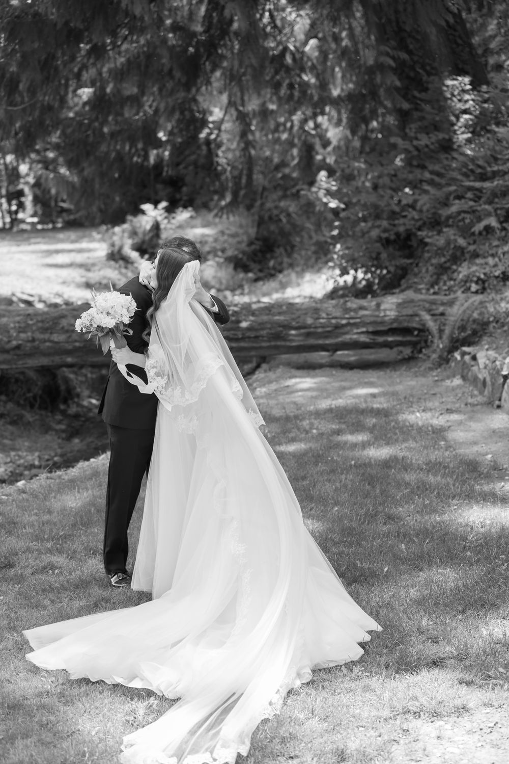 Black and white photo of bride in a long veil and flowing gown embraces groom in a dark suit in grassy forest. Acadia wedding photographer