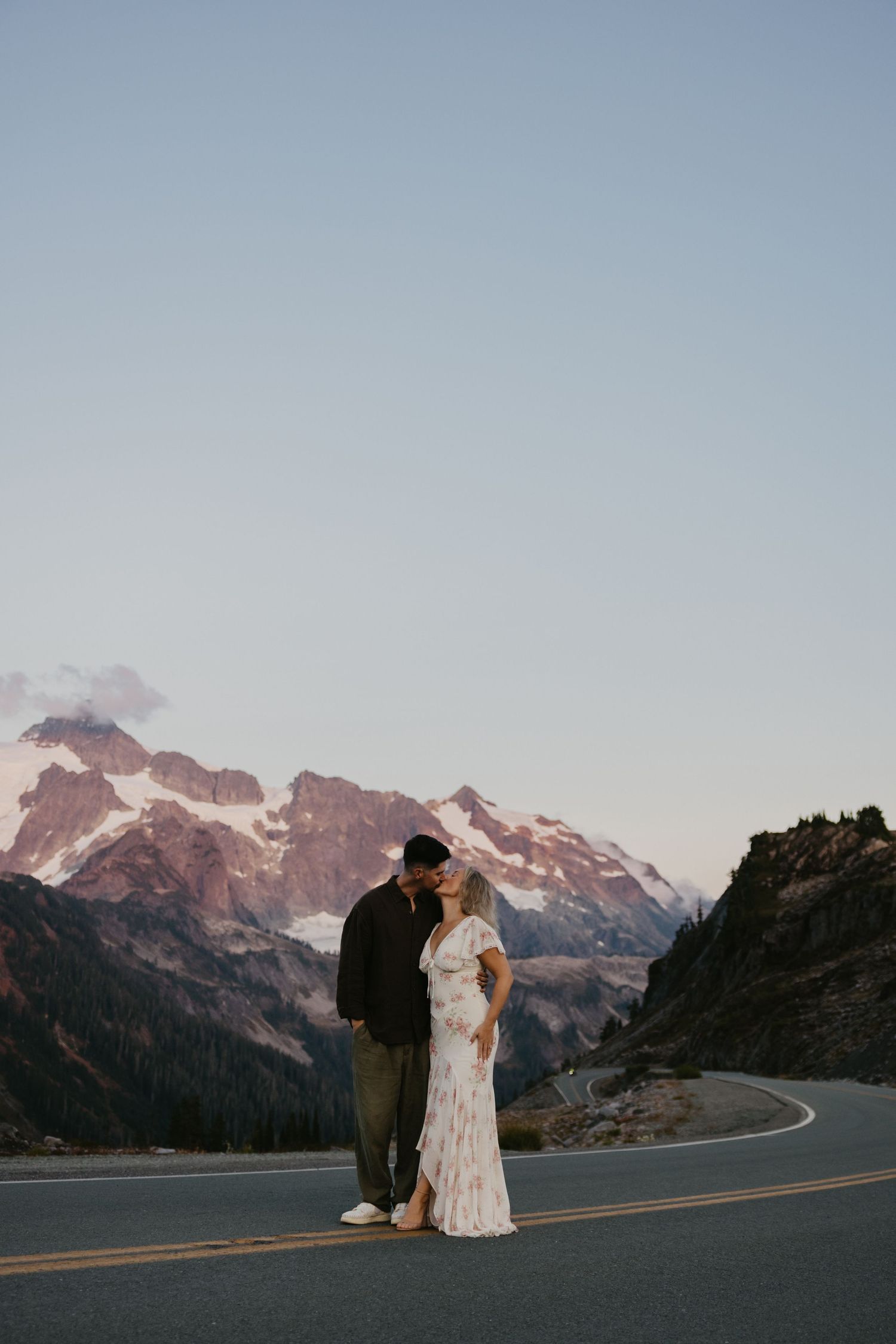 Couple kissing on a winding mountain road at dusk with snow-capped peaks in the background, an engagement session in the North Cascades