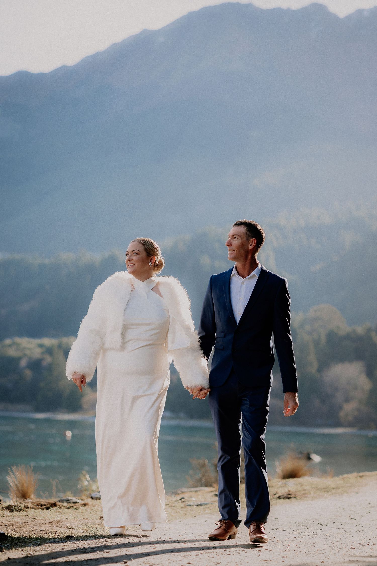 Bride in a white dress and fur shawl holds hands with a groom in a navy suit while walking along a lakeside path with mountains behind.