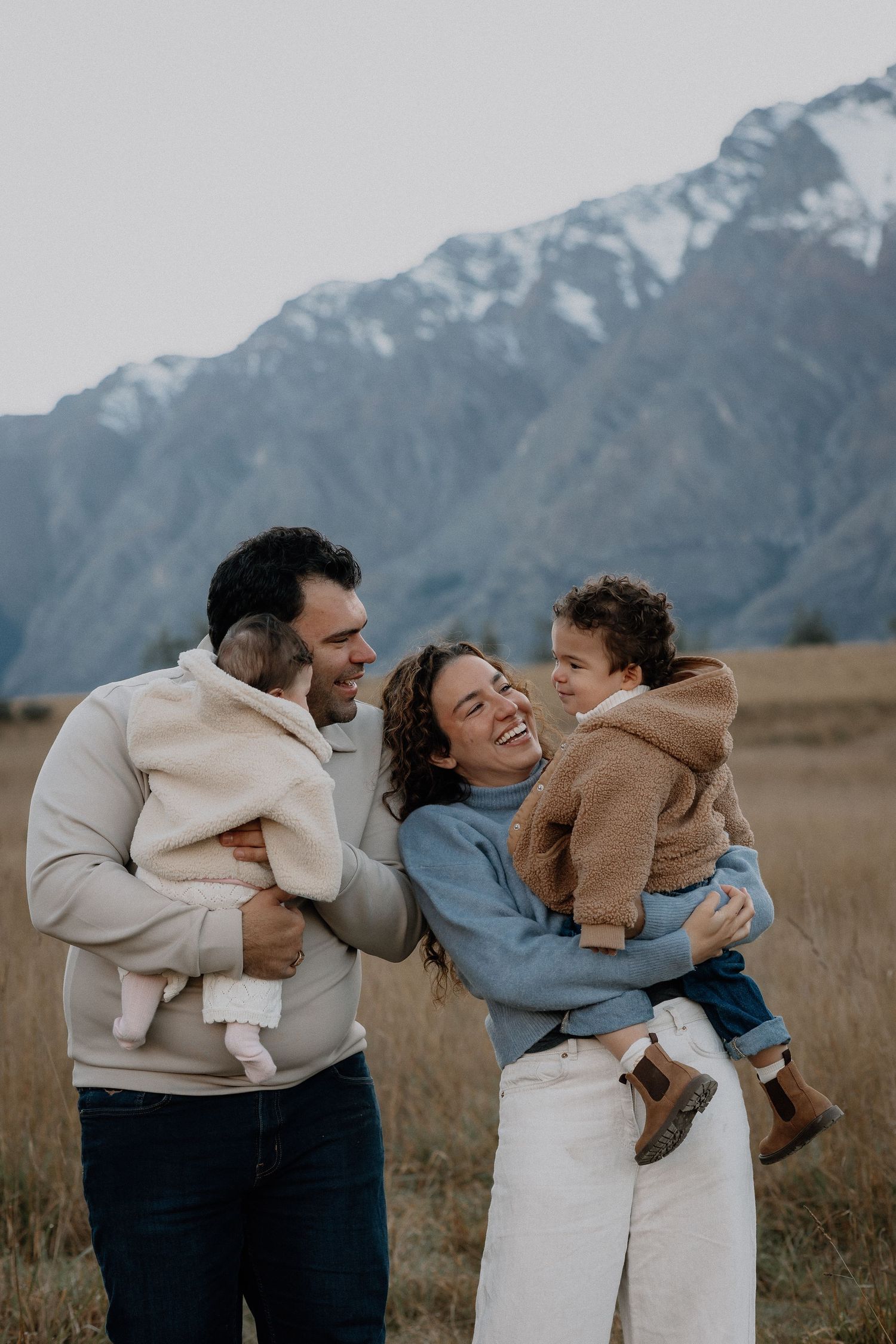A smiling family of four with two young children embraces in a golden grassy field with snow-capped mountains behind them.