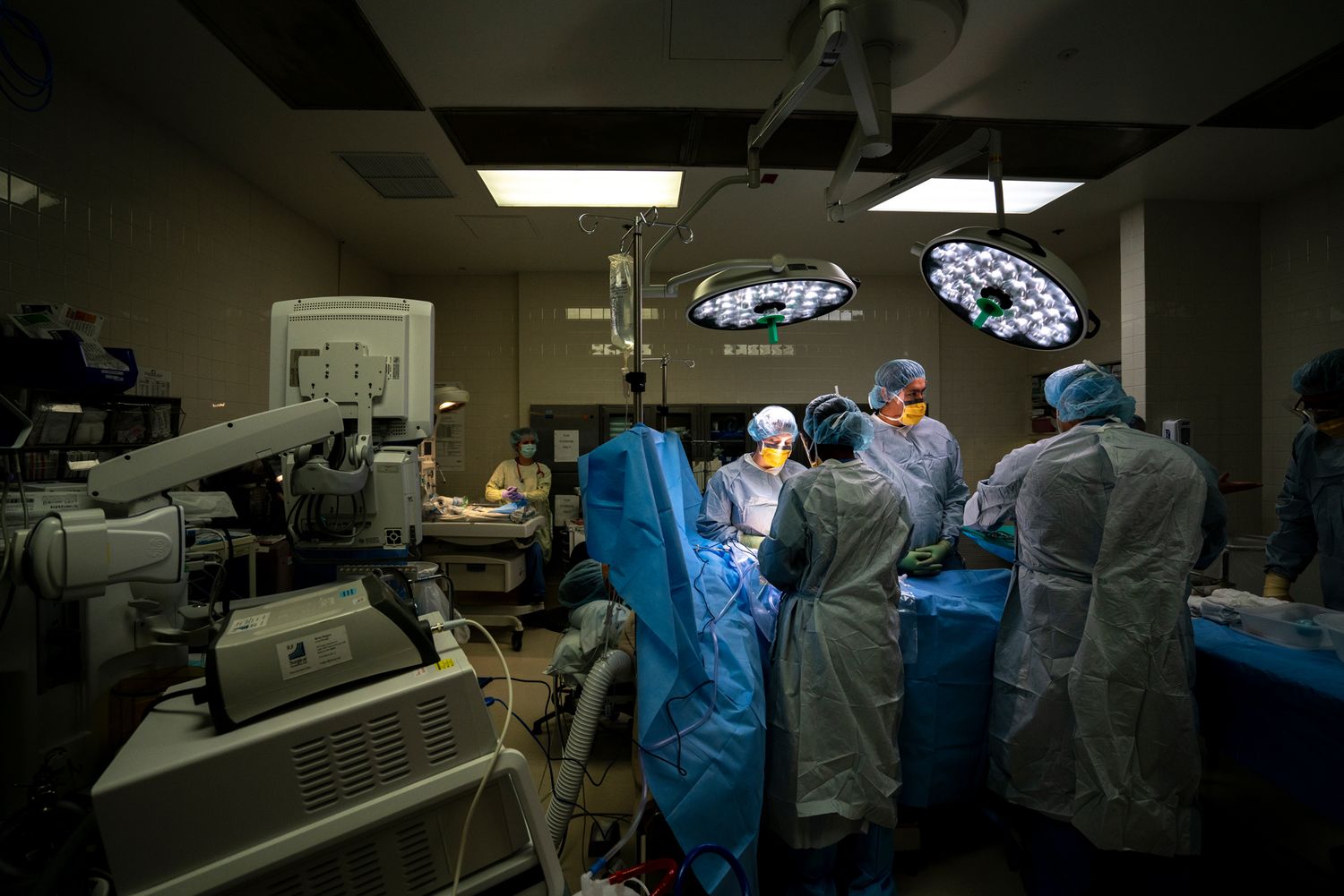 Surgical team in gowns, masks and caps operating under bright overhead lights in a dimly lit hospital operating room with medical equipment.