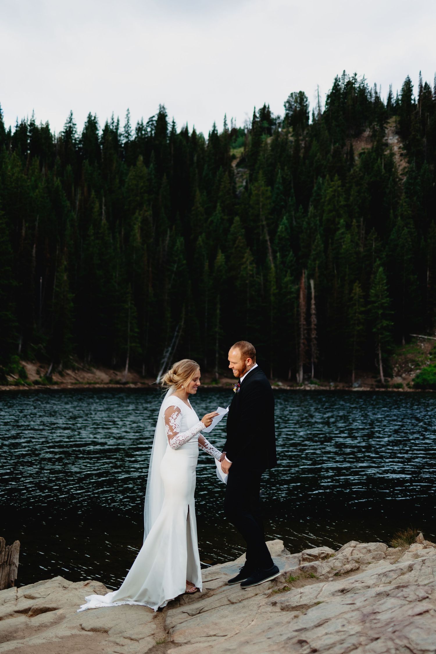 Bride in a long-sleeved white gown and veil reads vows to her groom on a rocky shore of a mountain lake framed by evergreen forest.