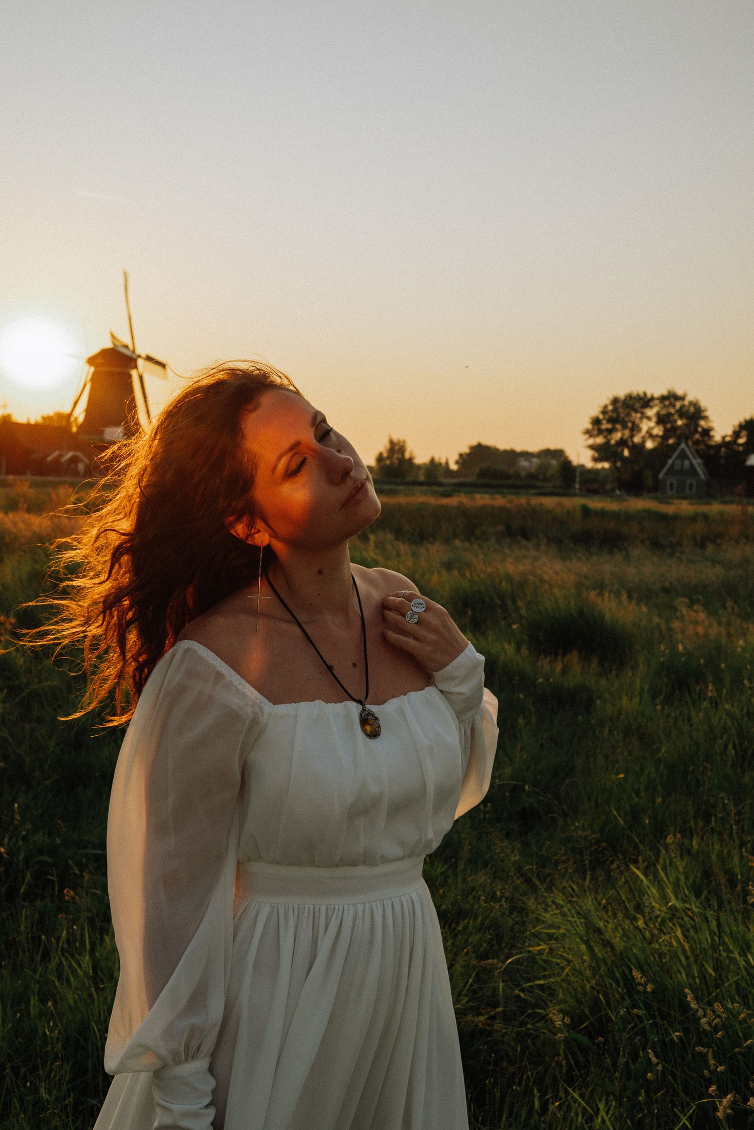 Woman in a white dress with curly hair stands in a grassy field at golden hour sunset with a windmill in the background, Zaanse Schans