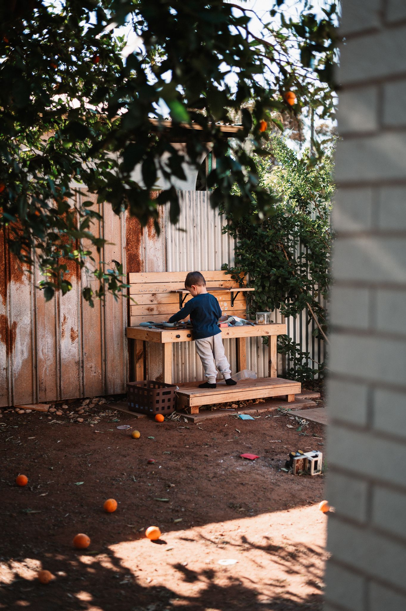 Young child in a navy sweater plays at a wooden outdoor mud kitchen in a sunlit backyard with fallen oranges scattered on the dirt.