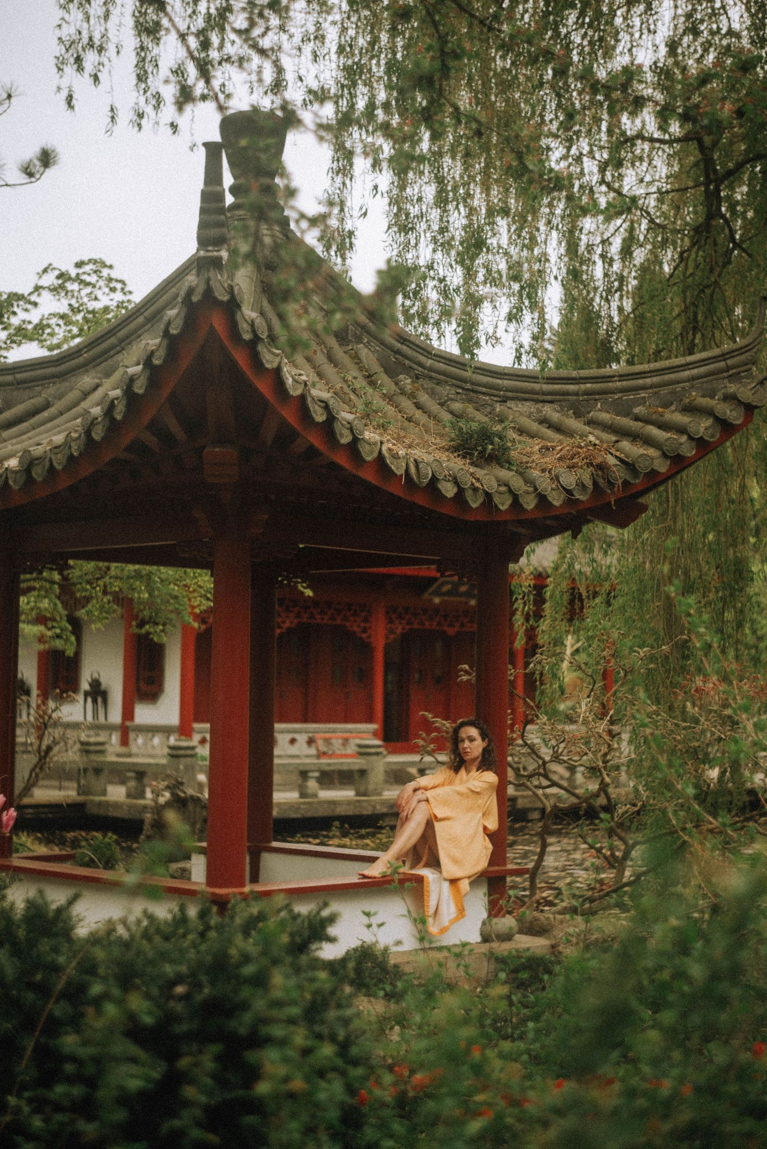 Woman in a yellow dress sits in Traditional pavilion at the pond in the Chinese garden at Hortus Haren.