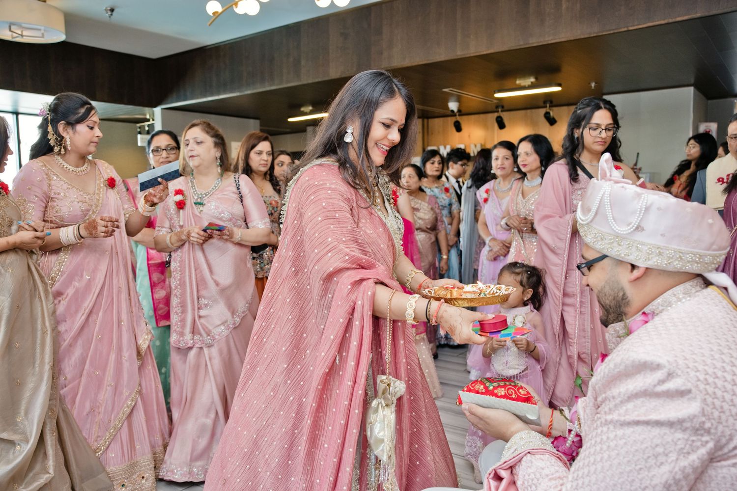 Woman in a pink embroidered sari holds a decorated ceremonial tray while guests in pink attire watch at an gujrati wedding celebration