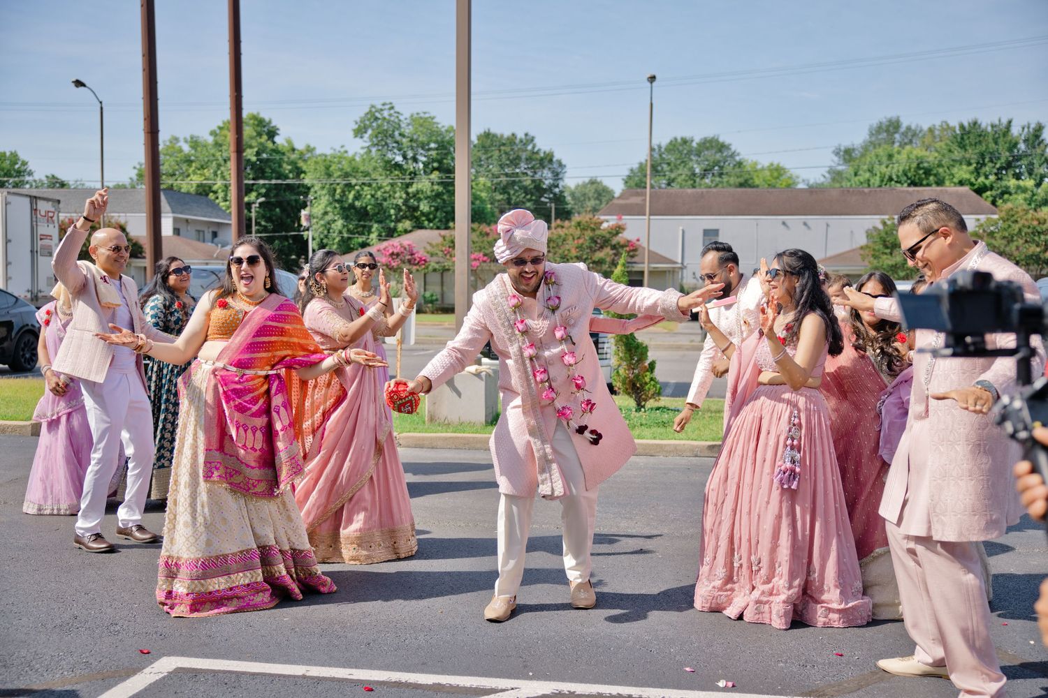 Groom in a white sherwani and pink turban dances with joyful guests in colorful Indian attire during an outdoor wedding baraat procession.