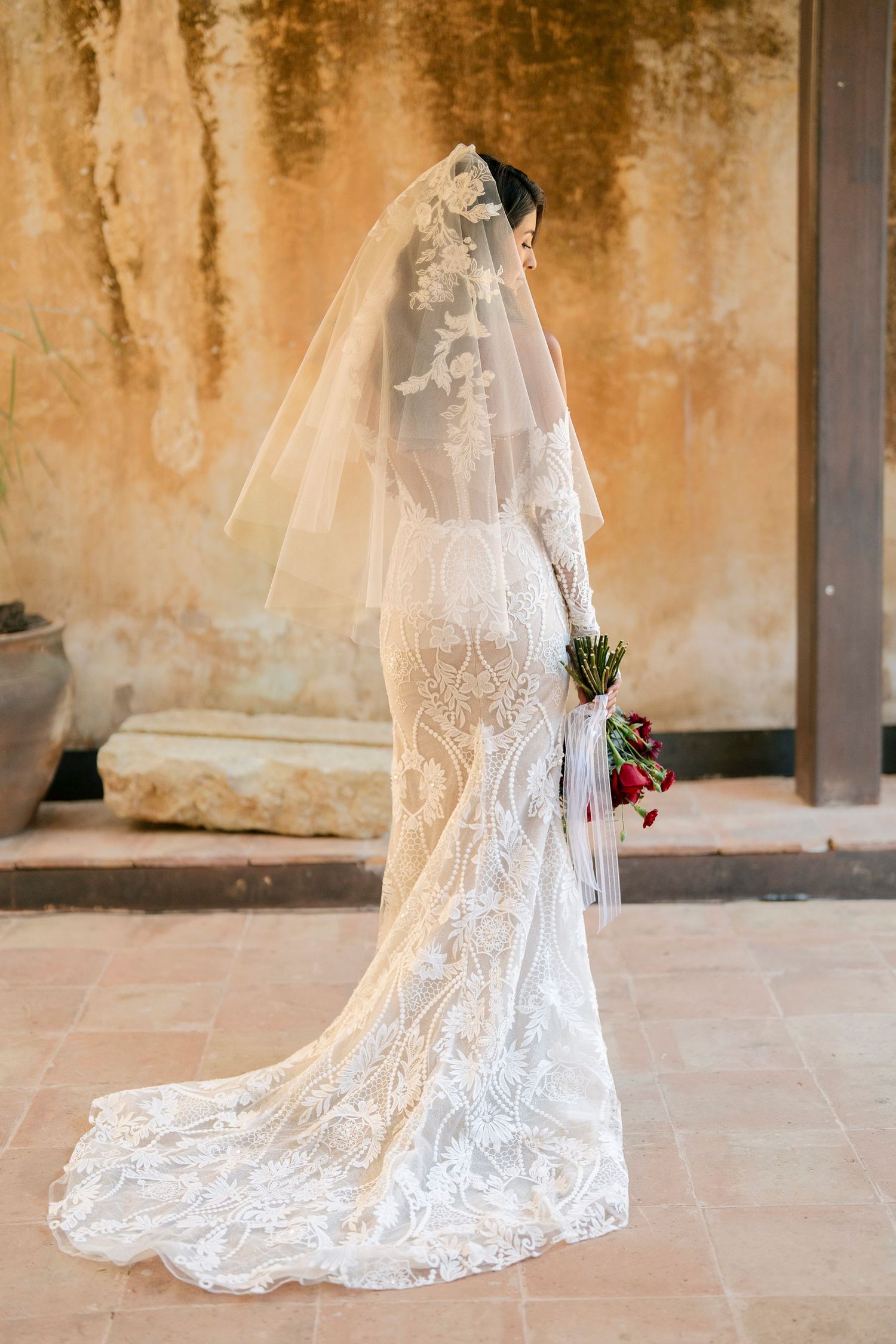 Bride in elegant lace wedding gown with long veil and floral bouquet standing against rustic wall.