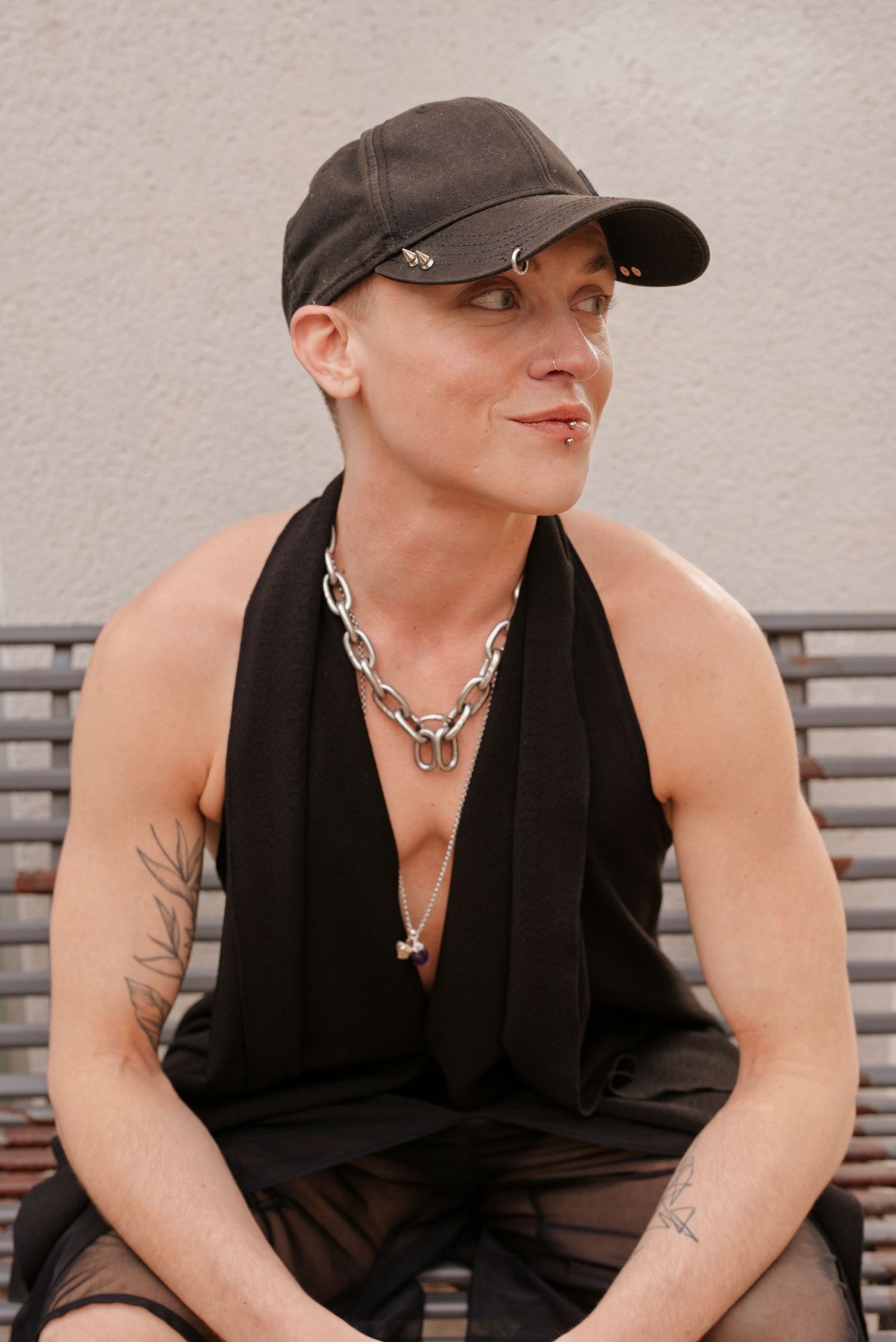 Person in black cap and halter top with chain necklace and tattoos sitting outdoors.