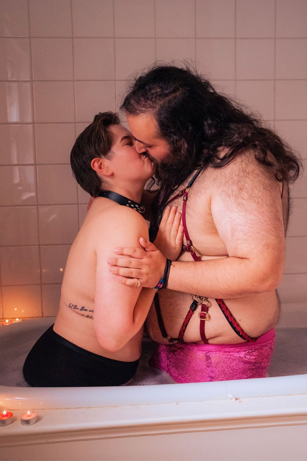 Two people sharing a romantic kiss in a candlelit bathtub, one wearing pink lingerie.