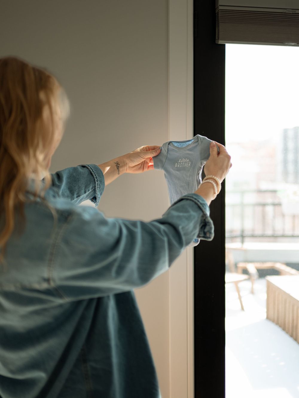 Woman in a denim shirt holds up a tiny light blue baby onesie near a sunlit window in a bright home.