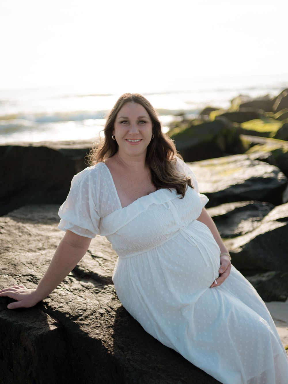 Smiling pregnant woman in a white dotted dress sitting on mossy rocks by the ocean during a golden hour maternity session.