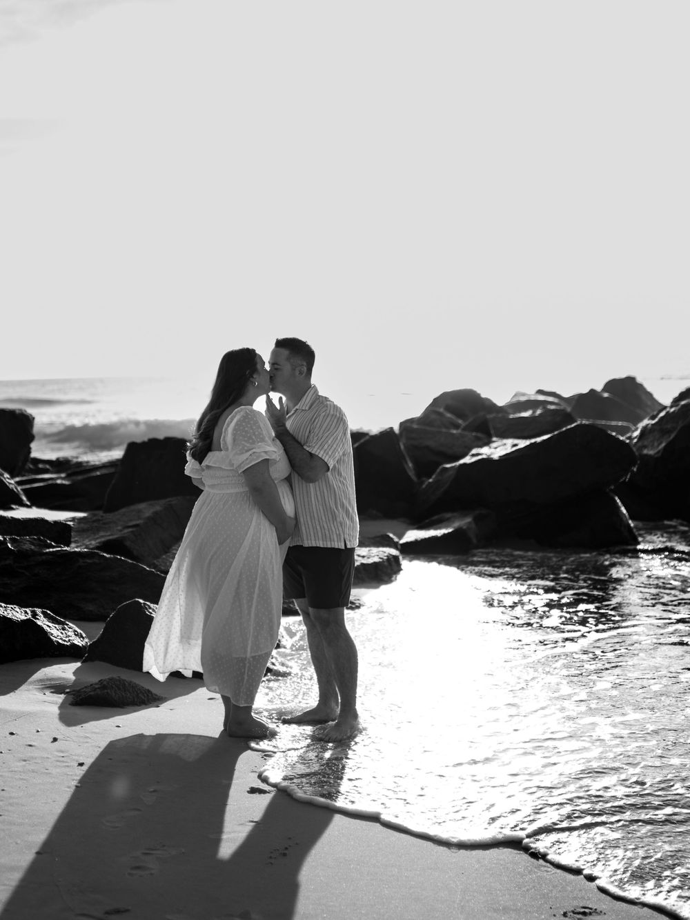 Black and white maternity photo of a couple kissing at the water's edge with the pregnant woman in a flowing dress near rocks.