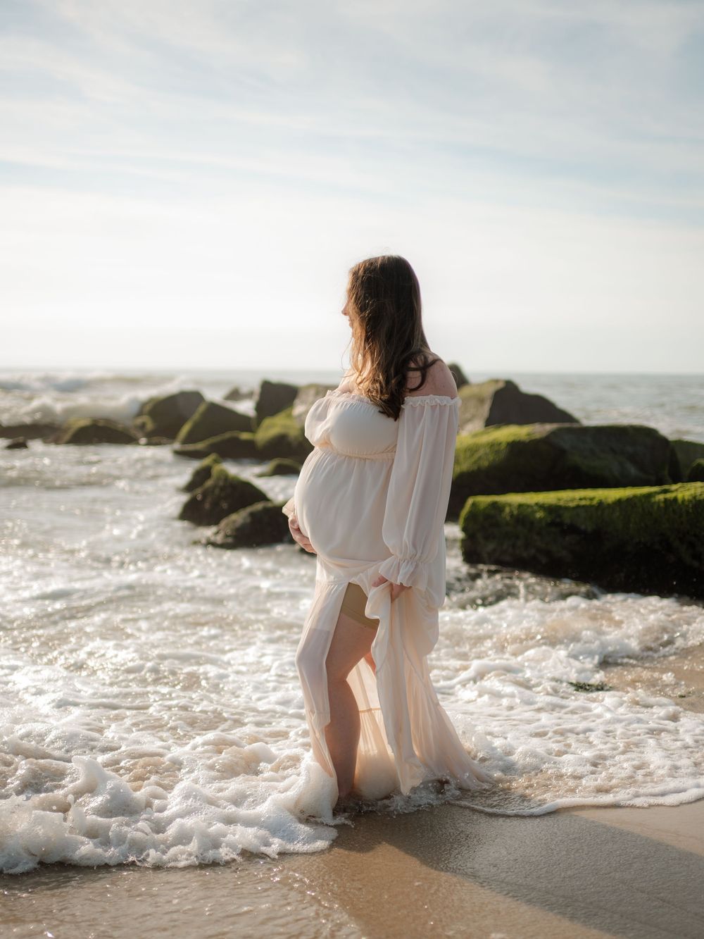 Pregnant woman in a flowing white off-shoulder gown standing in the ocean surf beside mossy rocks for a beach maternity photo.