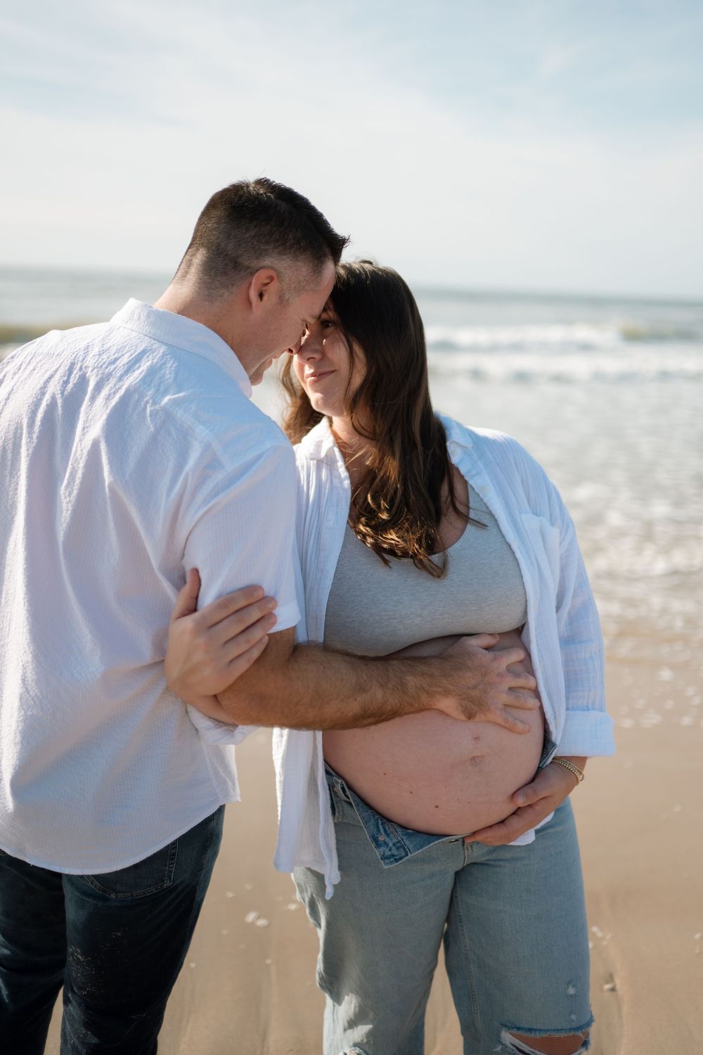 Couple embracing forehead to forehead on the beach, the man holding his pregnant partner's belly in a tender maternity portrait.