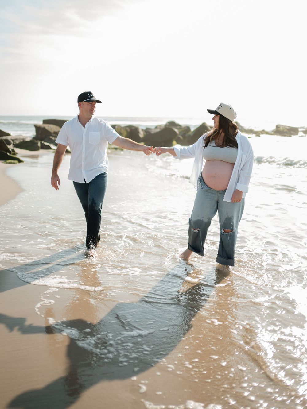 Expecting couple holding hands and walking barefoot through shallow ocean water, with the woman in a hat showing her baby bump.