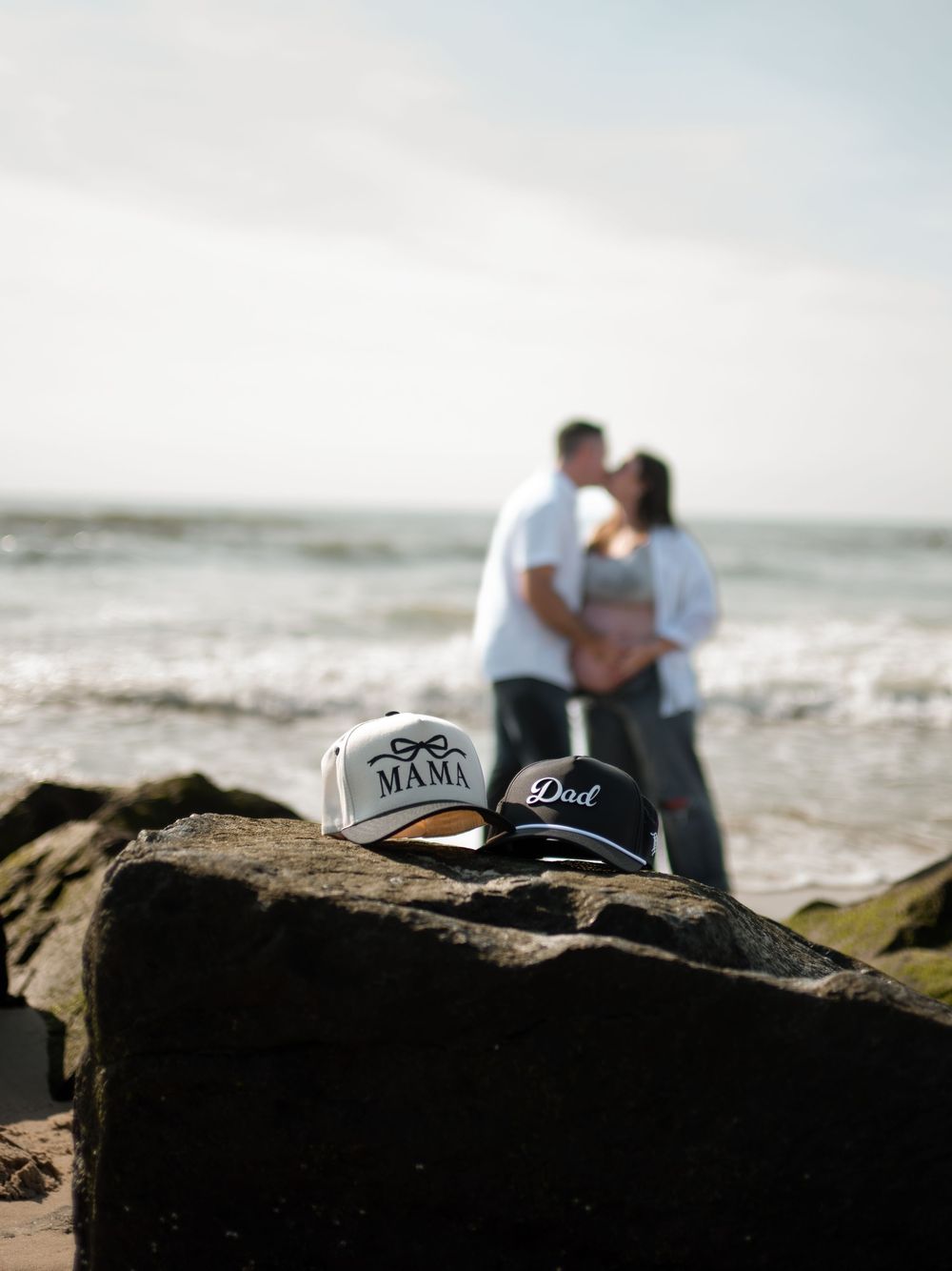 Mama and Dad baseball caps resting on a rock while a blurred couple kisses and cradles a baby bump on the beach behind.