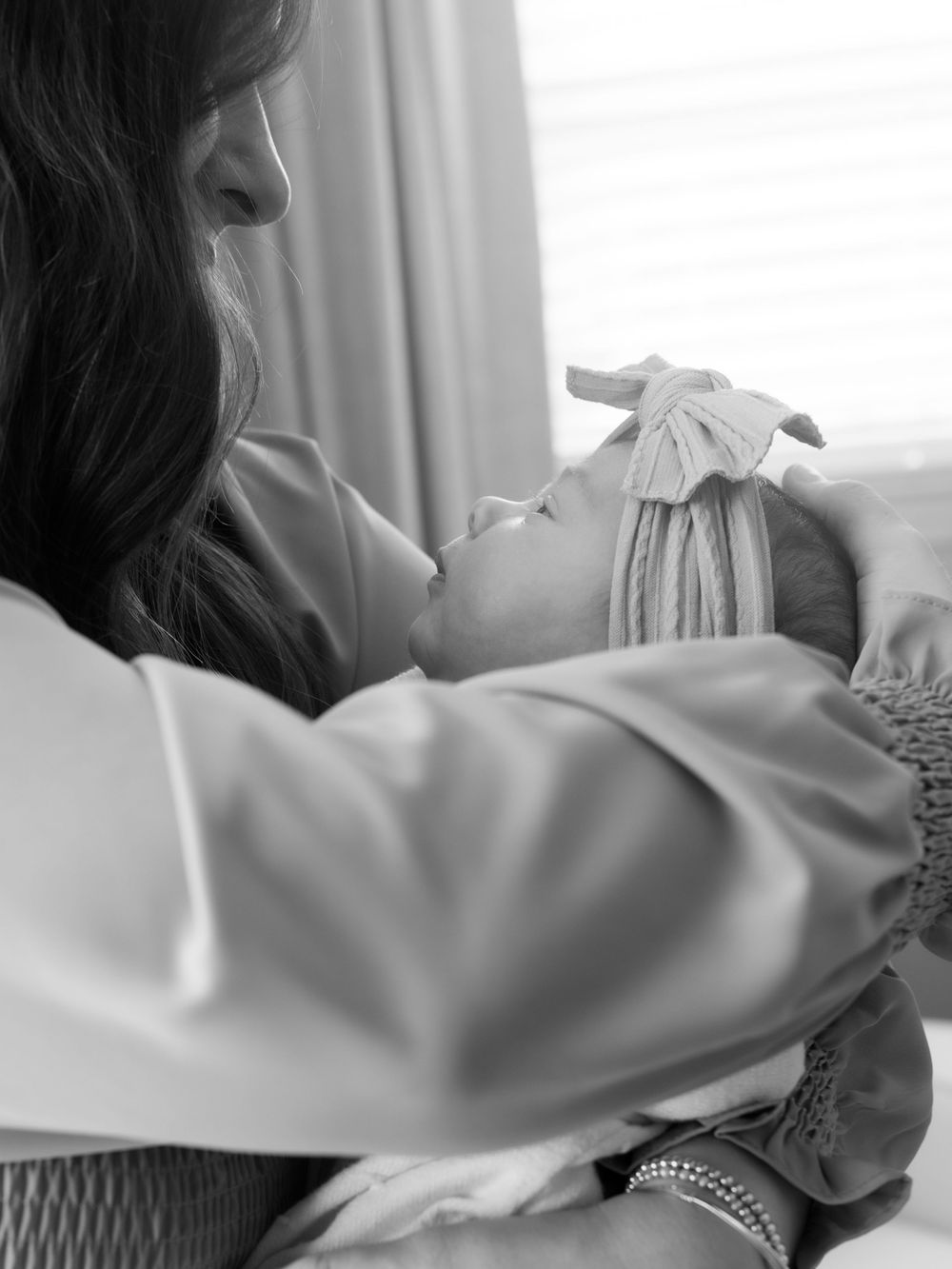 Black and white photo of a mother tenderly holding her newborn baby wearing a knotted bow headband.