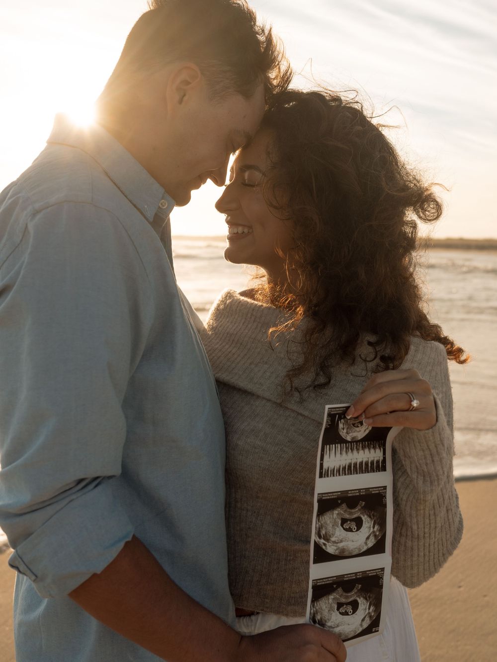 Couple touch foreheads on the beach at sunset as the woman holds ultrasound photos in a joyful pregnancy announcement.