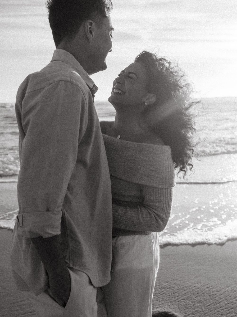 Black and white beach photo of a laughing woman with windswept curls embracing her partner by the ocean waves.