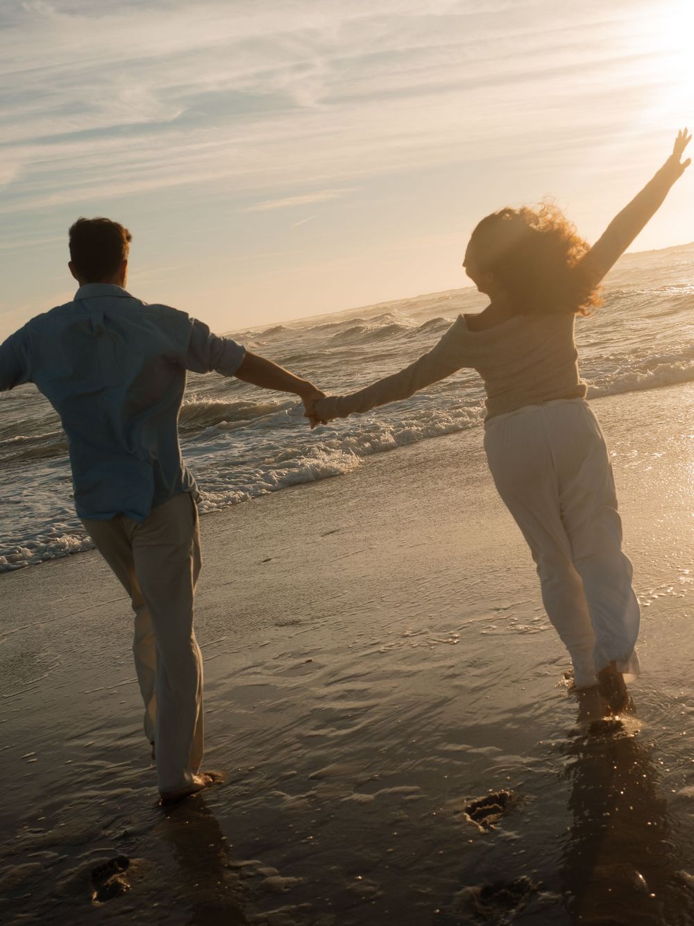Couple holds hands and runs along the shoreline at sunset, the woman raising one arm joyfully toward the glowing sky.
