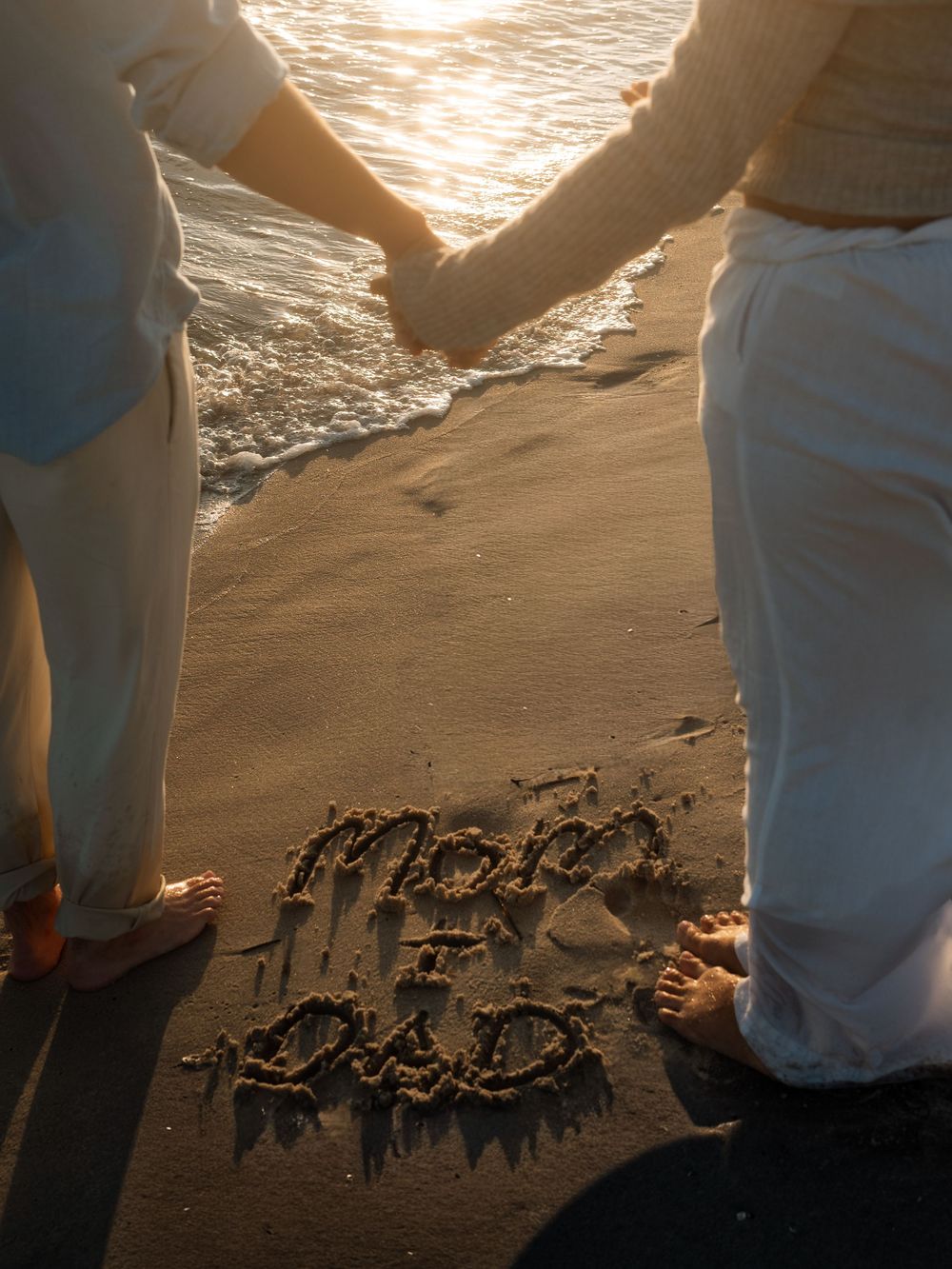 Couple holds hands on the beach beside Mom and Dad written in the sand as gentle waves roll in at golden hour.