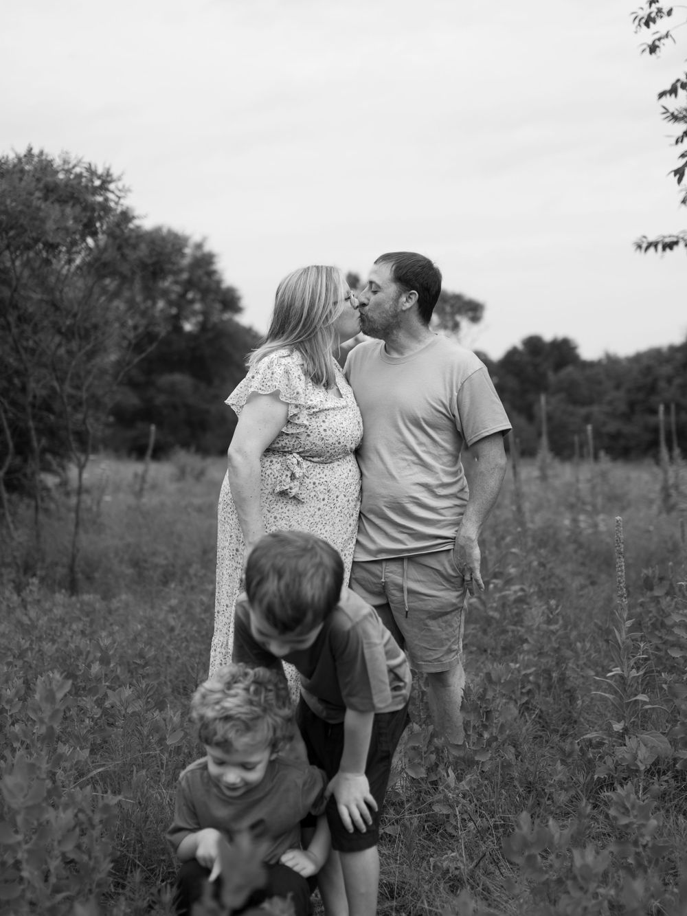 Black and white family portrait in a grassy field as a pregnant mother and father kiss while two young sons stand in front.