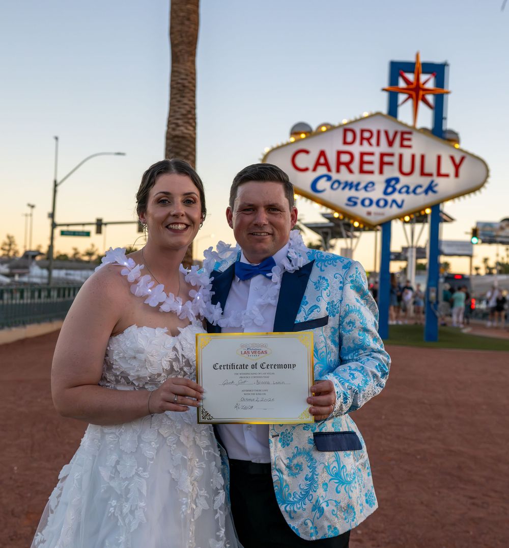 Smiling bride and groom in a floral jacket hold a Certificate of Ceremony in front of the Drive Carefully Come Back Soon Las Vegas sign.