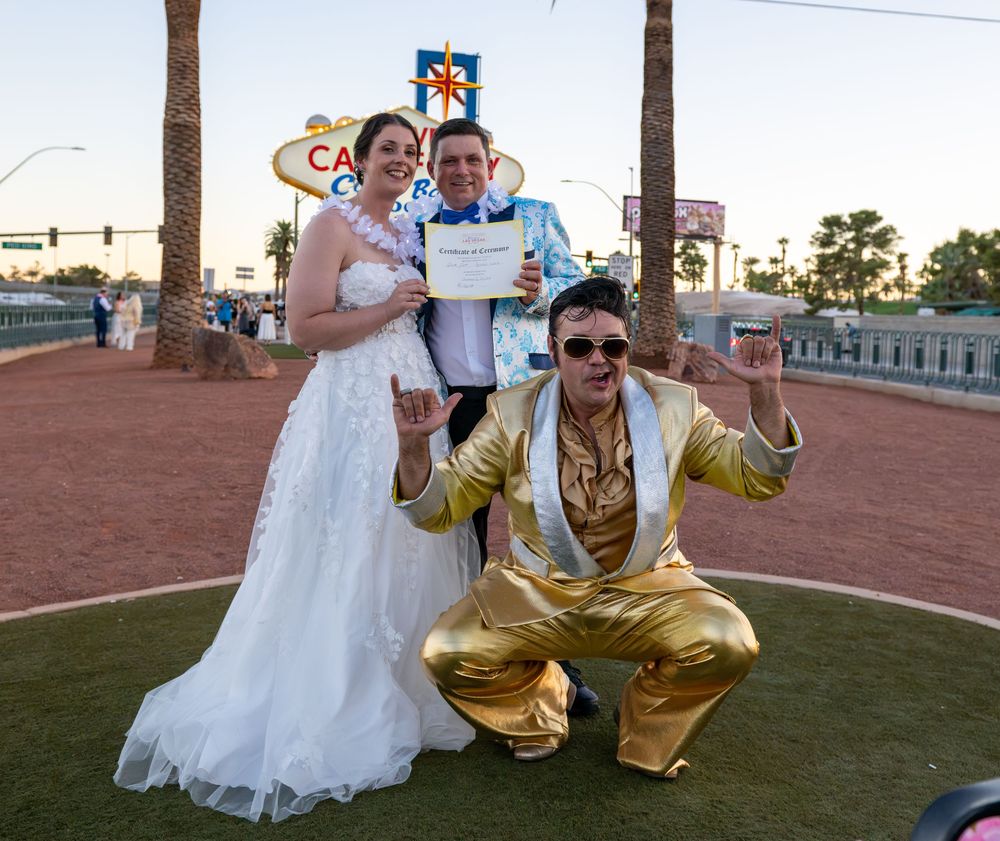 Bride in white gown and officiant hold a marriage certificate beside an Elvis impersonator in gold at the Welcome to Las Vegas sign.