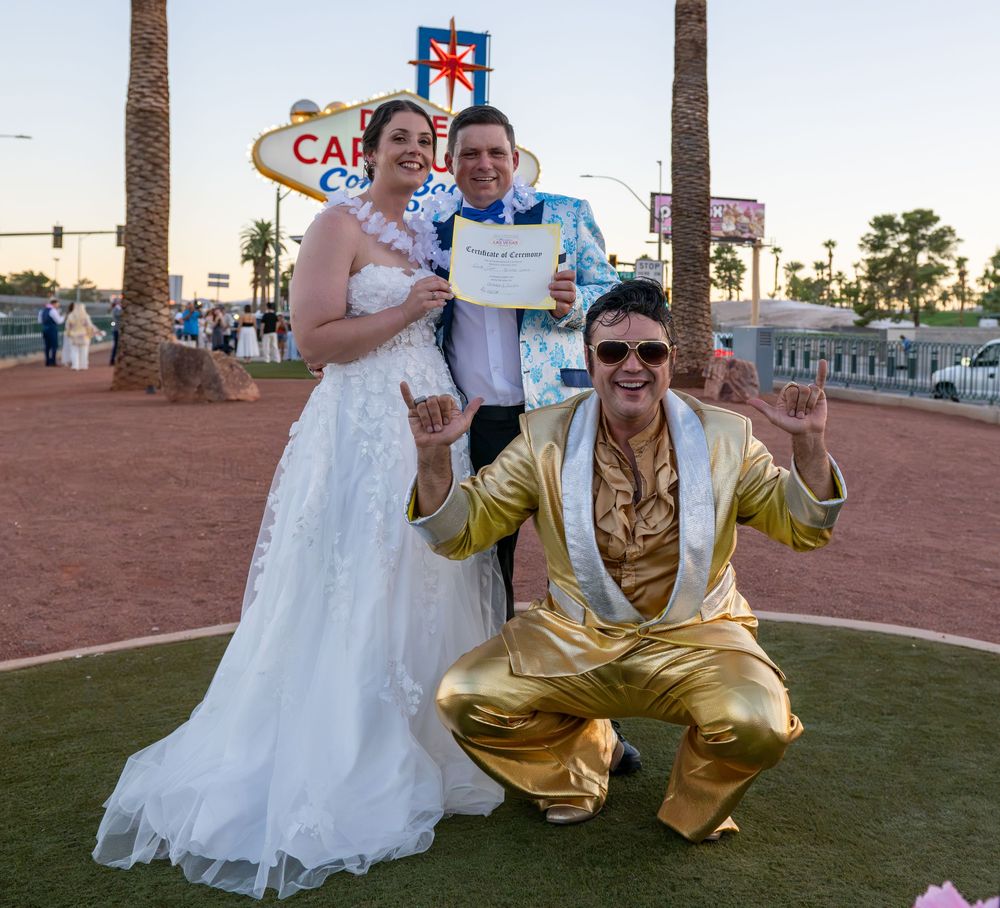 Bride in a white gown and groom hold a marriage certificate near a Las Vegas sign while an Elvis impersonator in a gold suit poses.
