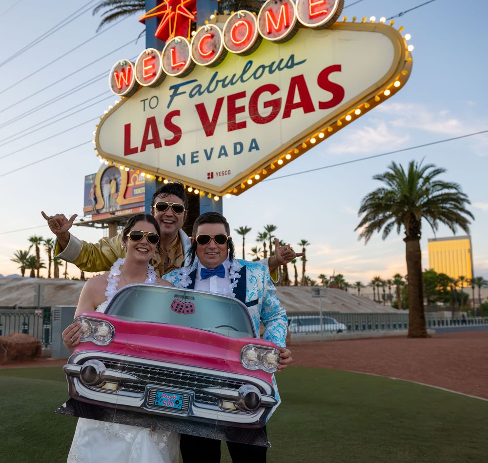 Newlywed couple in sunglasses pose with a pink Cadillac prop and an Elvis impersonator at the Welcome to Fabulous Las Vegas sign.