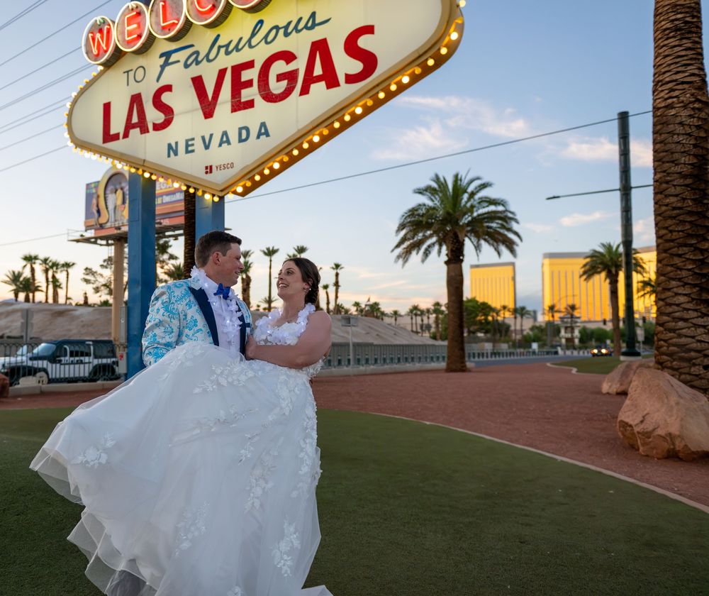 Newlyweds embrace on the grass in front of the Welcome to Fabulous Las Vegas sign with palm trees and a flowing white wedding dress.