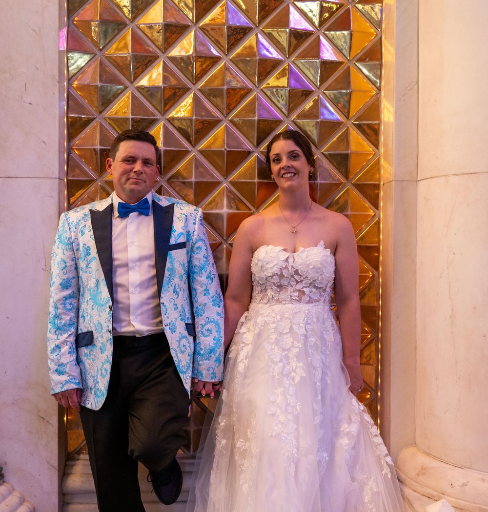 A smiling bride in a strapless lace wedding gown stands holding hands with a groom in a blue floral tuxedo jacket and bow tie.