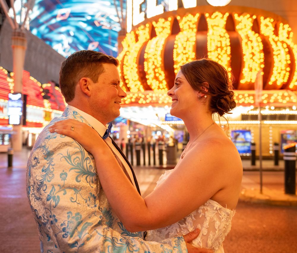 A smiling couple embraces in front of glowing neon lights on a lively Las Vegas street at night, he in a floral blazer, she in a lace dress.