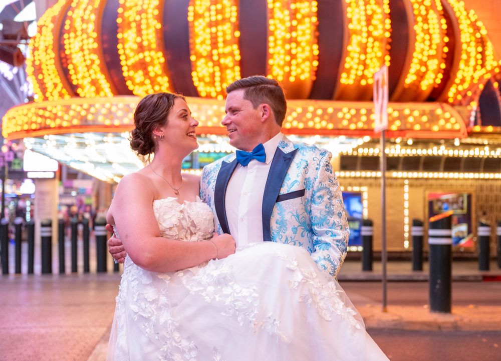 Smiling groom in a turquoise floral tuxedo jacket holds his bride in a white lace gown beneath glowing marquee lights on a Las Vegas street.