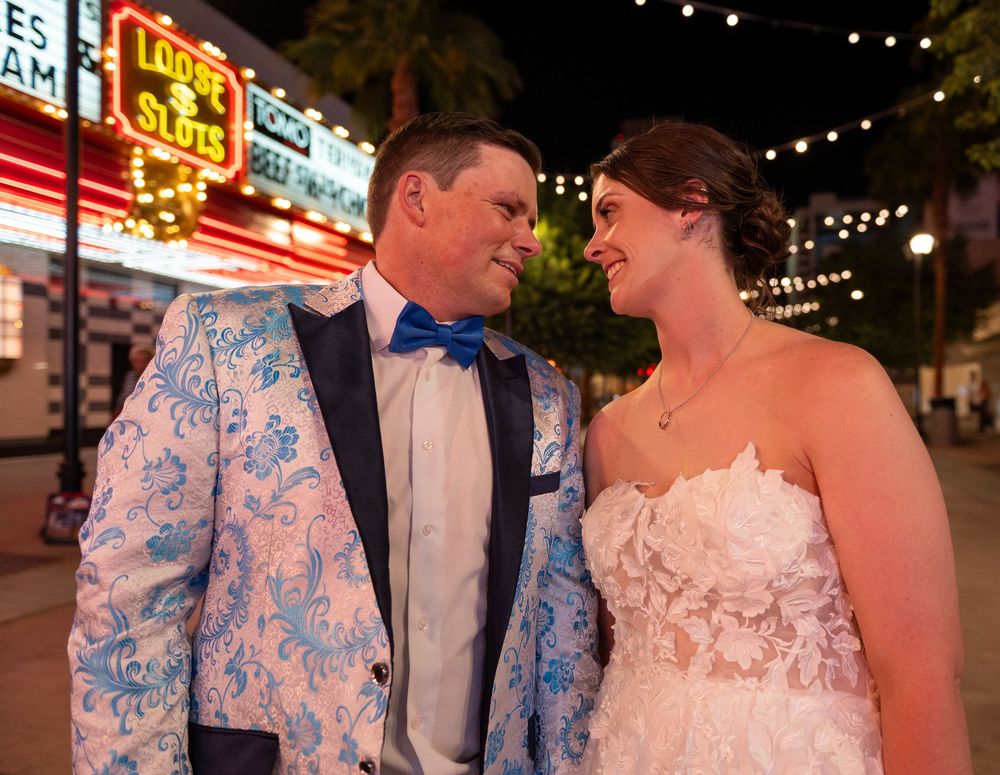 Bride in a lace wedding gown smiles at a groom in a blue floral tuxedo jacket and bow tie on a neon-lit Las Vegas street at night.