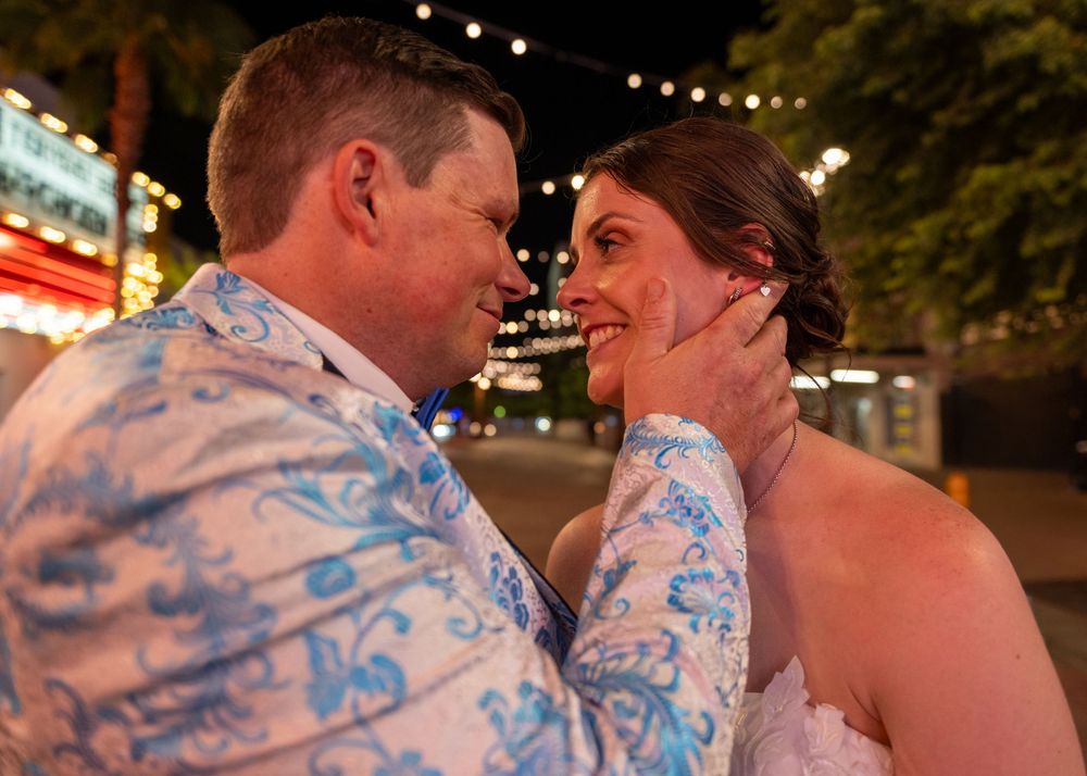 Smiling couple share a tender moment at night, the man in a blue floral jacket touching the woman's face, with glowing city lights behind.