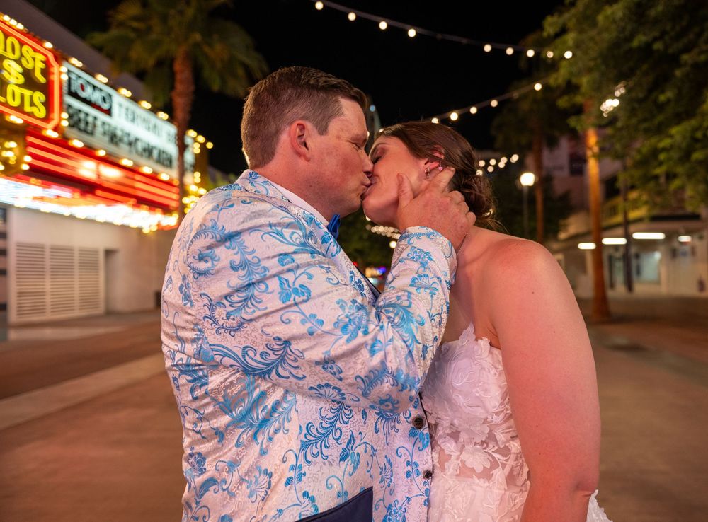 A bride in a white lace gown and a groom in a blue floral jacket kiss on a Las Vegas street at night.