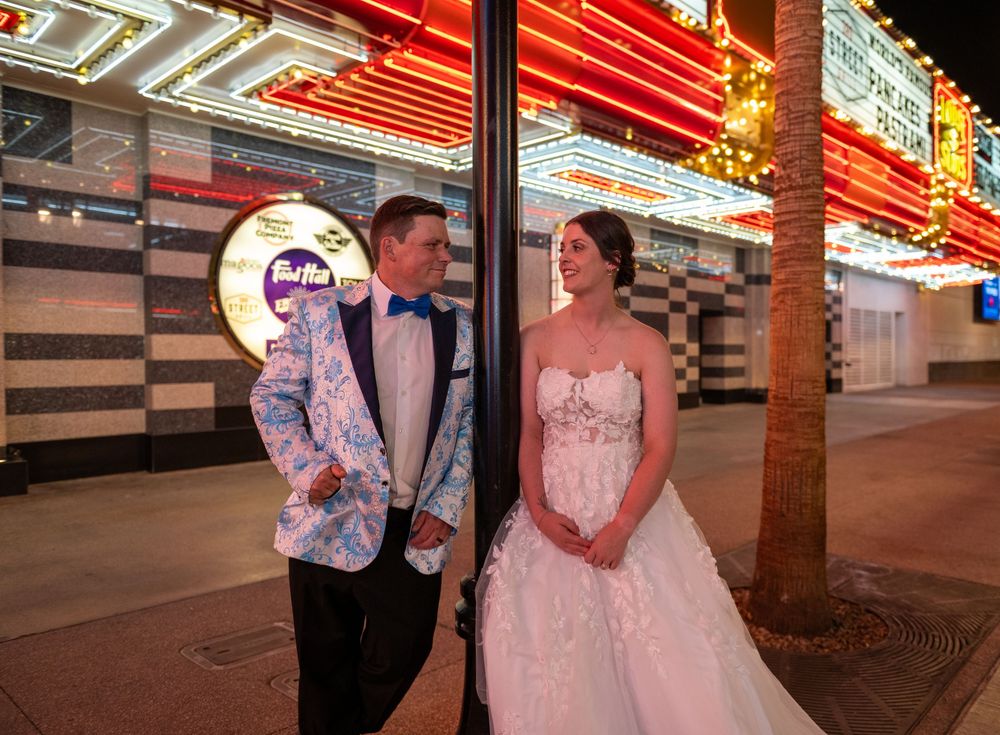 Bride in a strapless lace wedding gown and groom in a floral blazer pose together on a Las Vegas street under glowing neon lights.