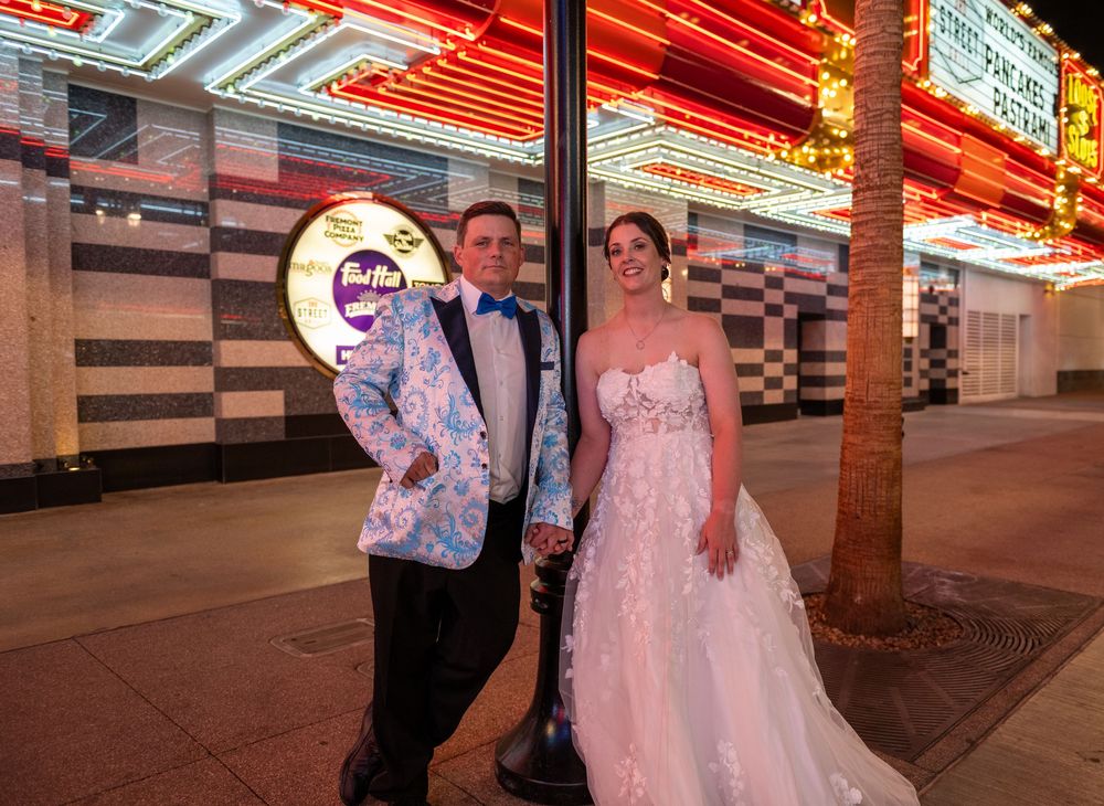 Bride in a white lace gown and groom in a floral blue jacket pose hand in hand beneath glowing neon lights on a Las Vegas street.