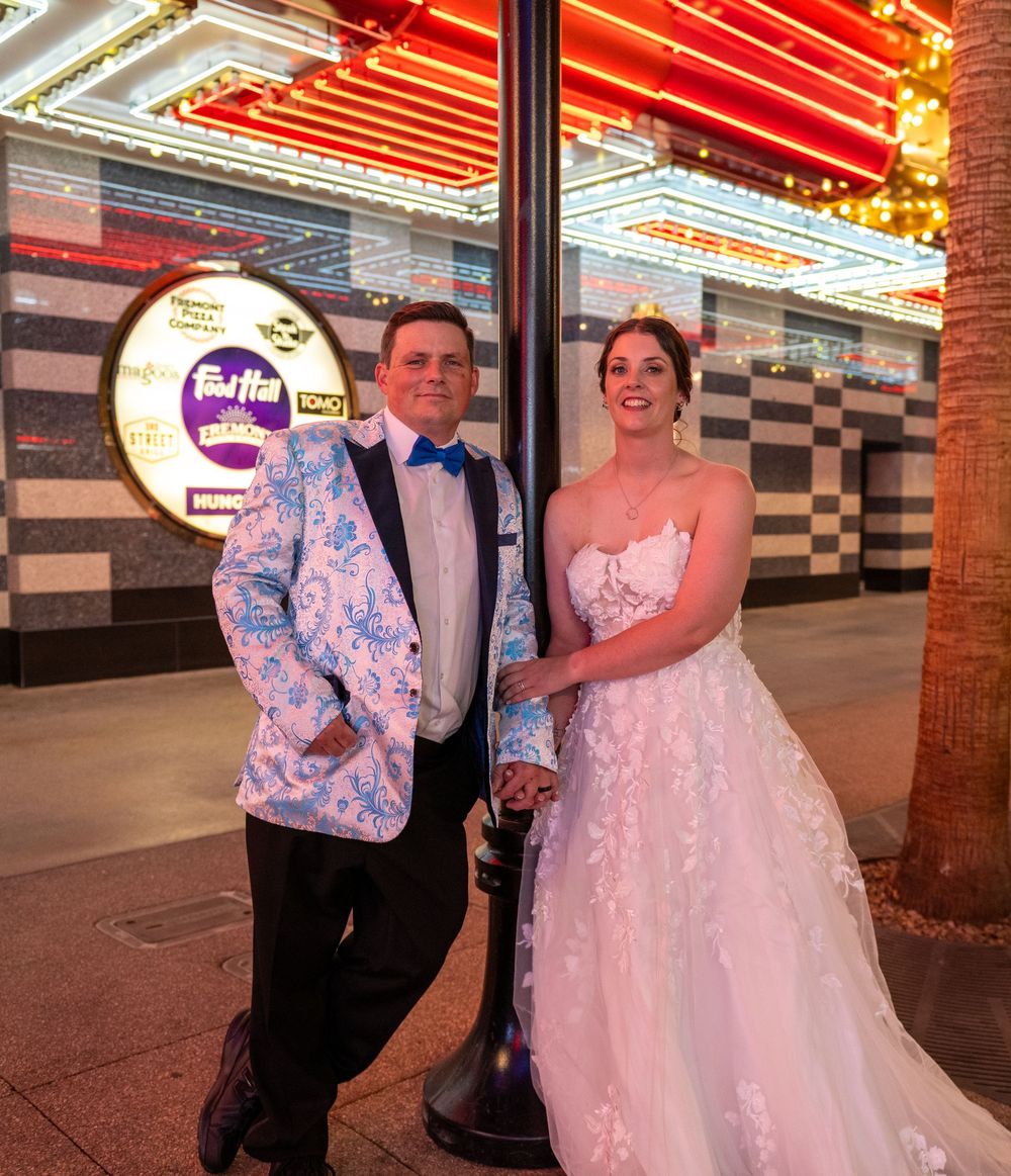 Bride in a lace gown and groom in a blue floral tuxedo jacket pose by a lamppost under glowing Las Vegas neon lights.