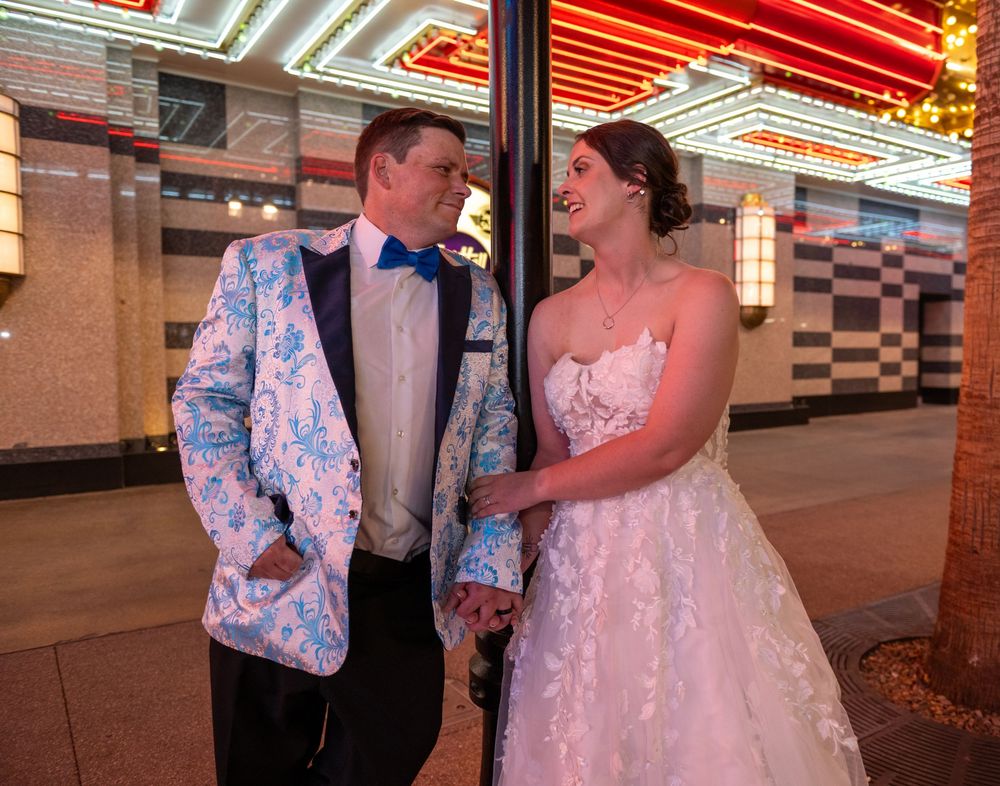 Bride in a lace wedding gown smiles at her groom in a blue floral tuxedo jacket beside a pole on a neon-lit Las Vegas street.