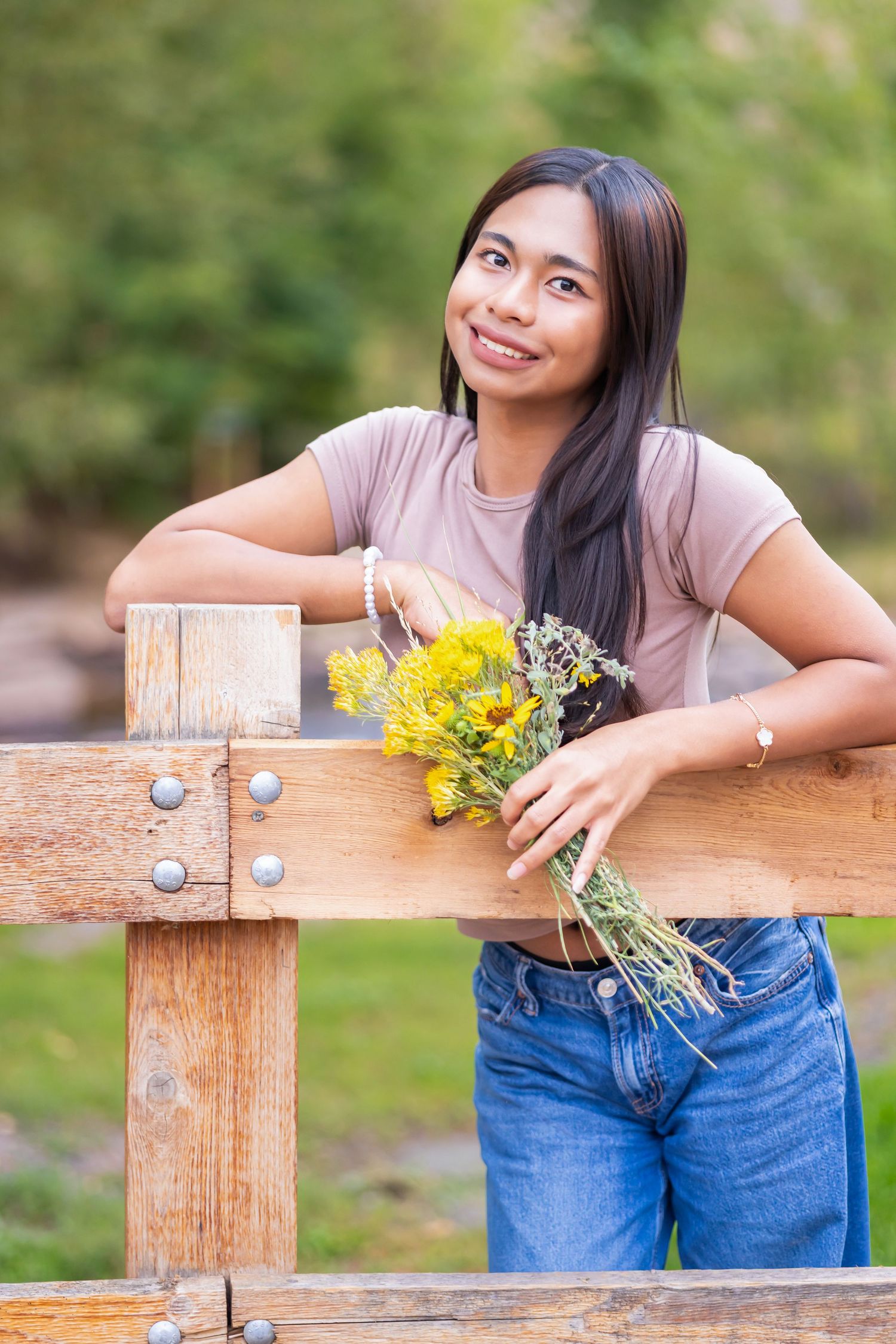 Young Asian woman smiling, leaning on wooden fence holding yellow wildflowers, wearing jeans and beige top outdoors.