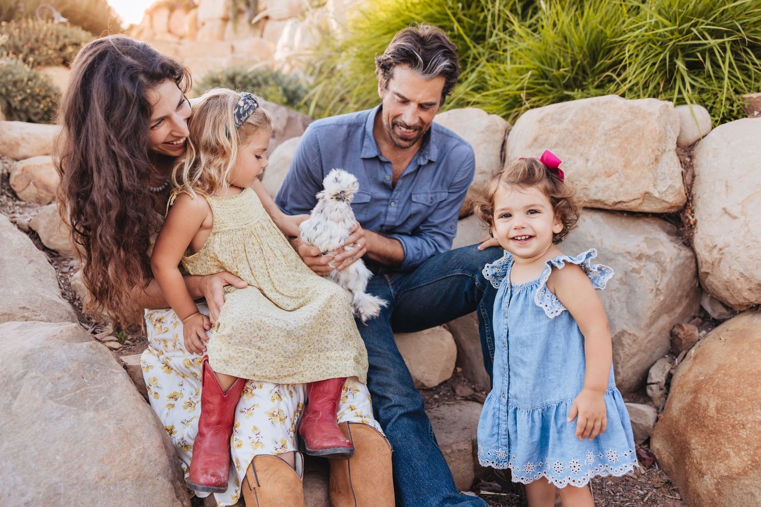 A smiling family with two young girls and a small white puppy sit together on large sandstone rocks outdoors in warm golden sunlight.